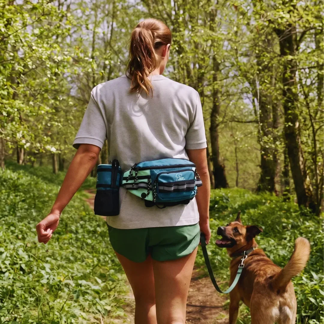 A woman on a woodland trail wearing the hip pack with the sling bag attached, her dog walking beside her on a teal leash