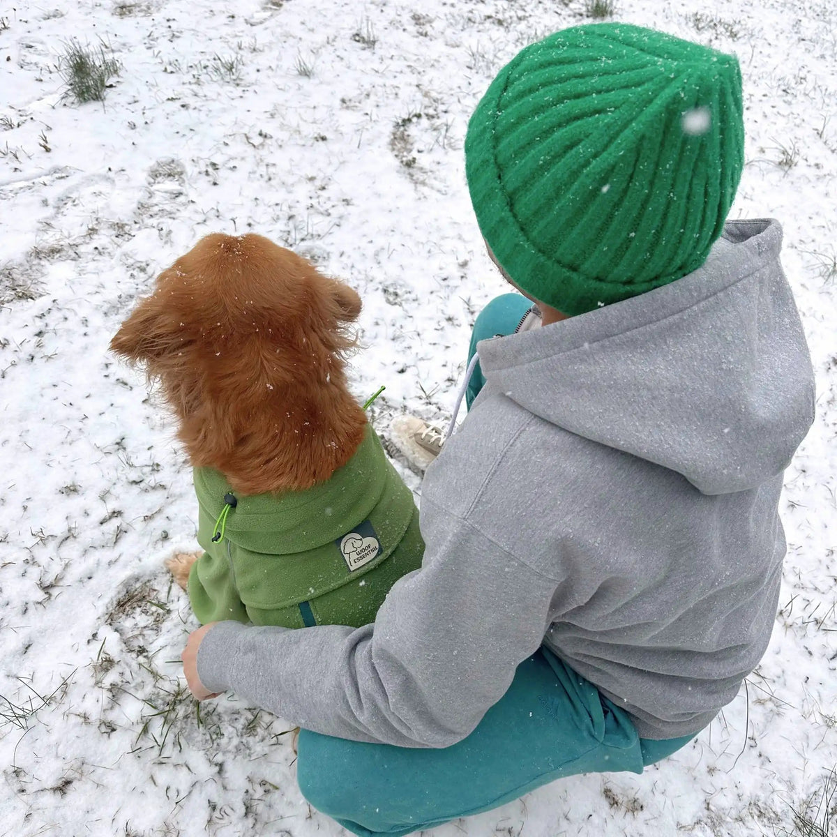 A woman in a green beanie sitting in the snow with her dog in the matching green fleece