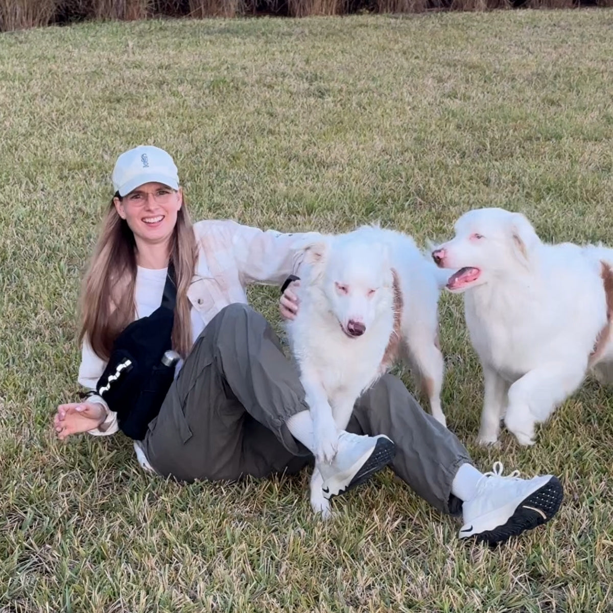Sitting in the grass with two white dogs, black Hip Pack 2.0 worn crossbody